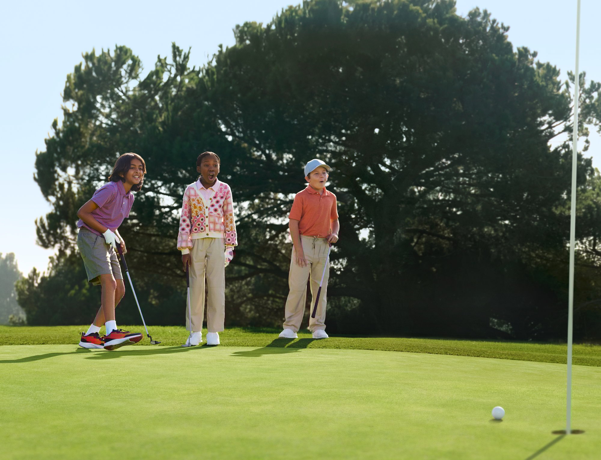 Three happy youth golfers stand on a golf course, clubs in hand, looking surprised by something in the distance