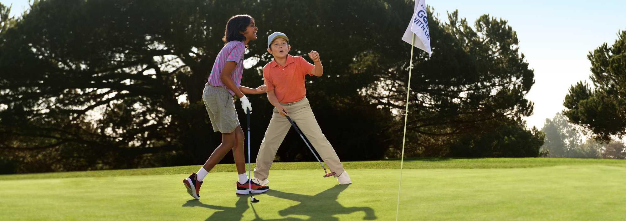 Two youth golfers celebrate next to the hole and a Golf with Us flag