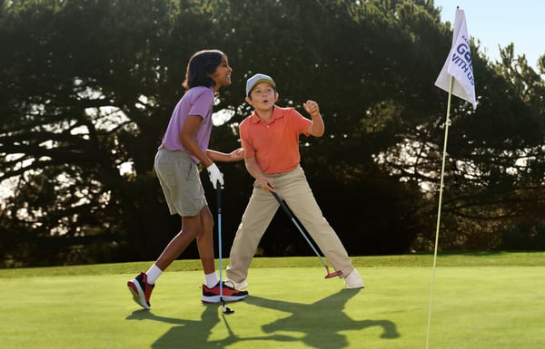 Two youth golfers celebrate next to the hole and a Golf with Us flag