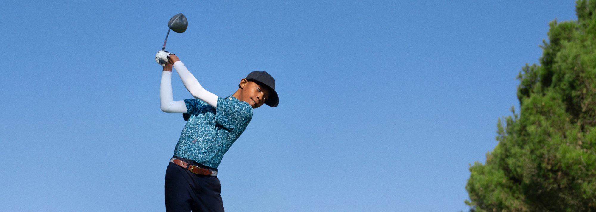 A child in a blue hat and a patterned shirt swings a club on a golf course.