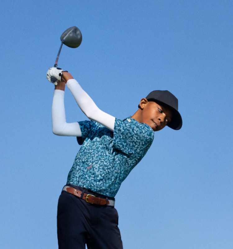 A child in a blue hat and a patterned shirt swings a club on a golf course.