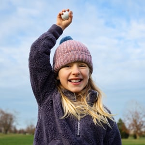 A young girl in a knitted hat smiles while holding a golf ball above her head.