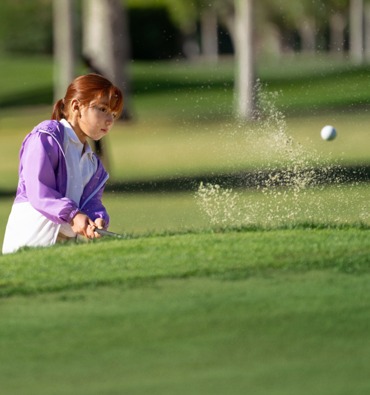 A Girl in a purple windbreaker hits a golf ball out of a sand trap.