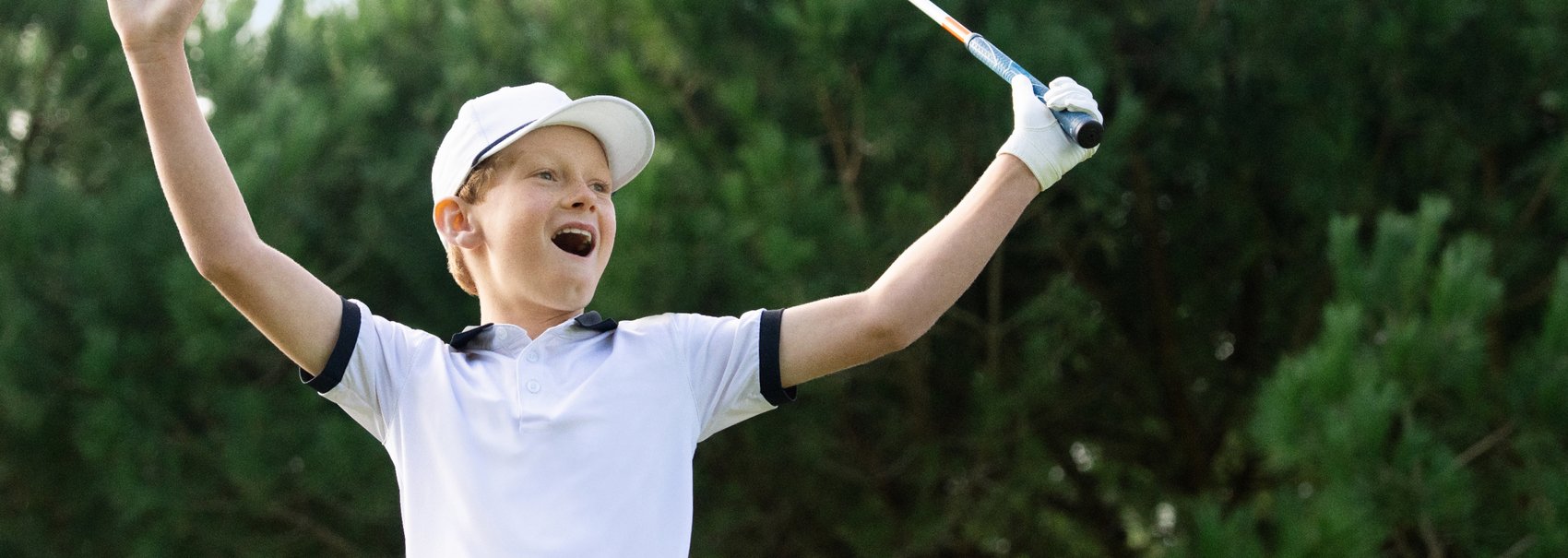 A boy in a white hat and a white collared shirt holds a golf club as he raises his arms in the air.