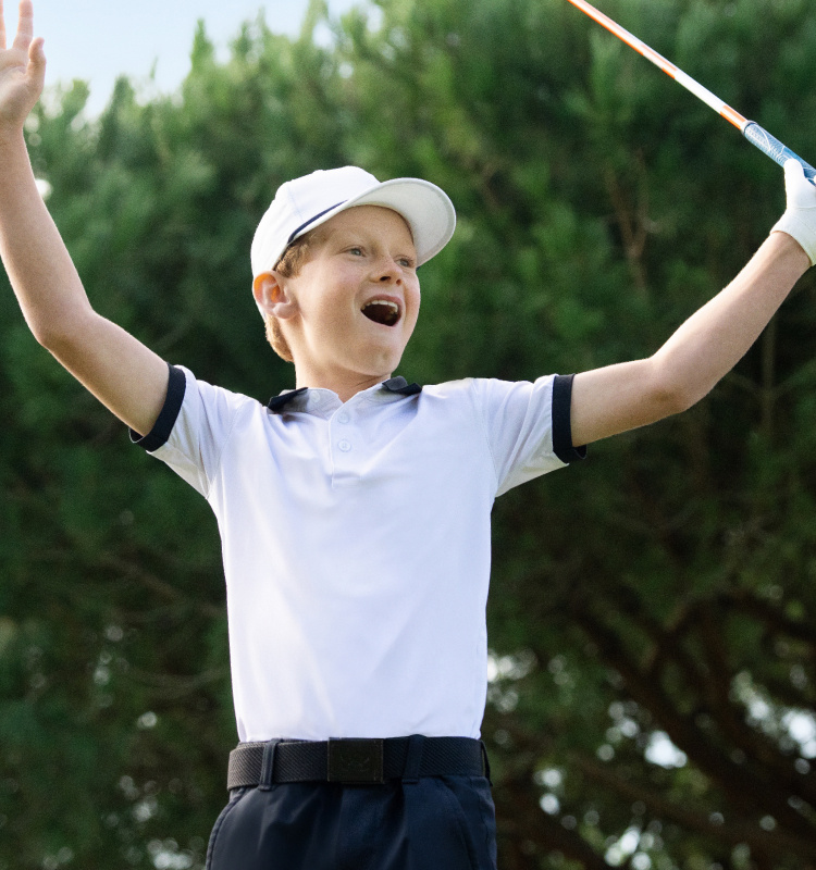 A boy in a white hat and a white collared shirt holds a golf club as he raises his arms in the air.